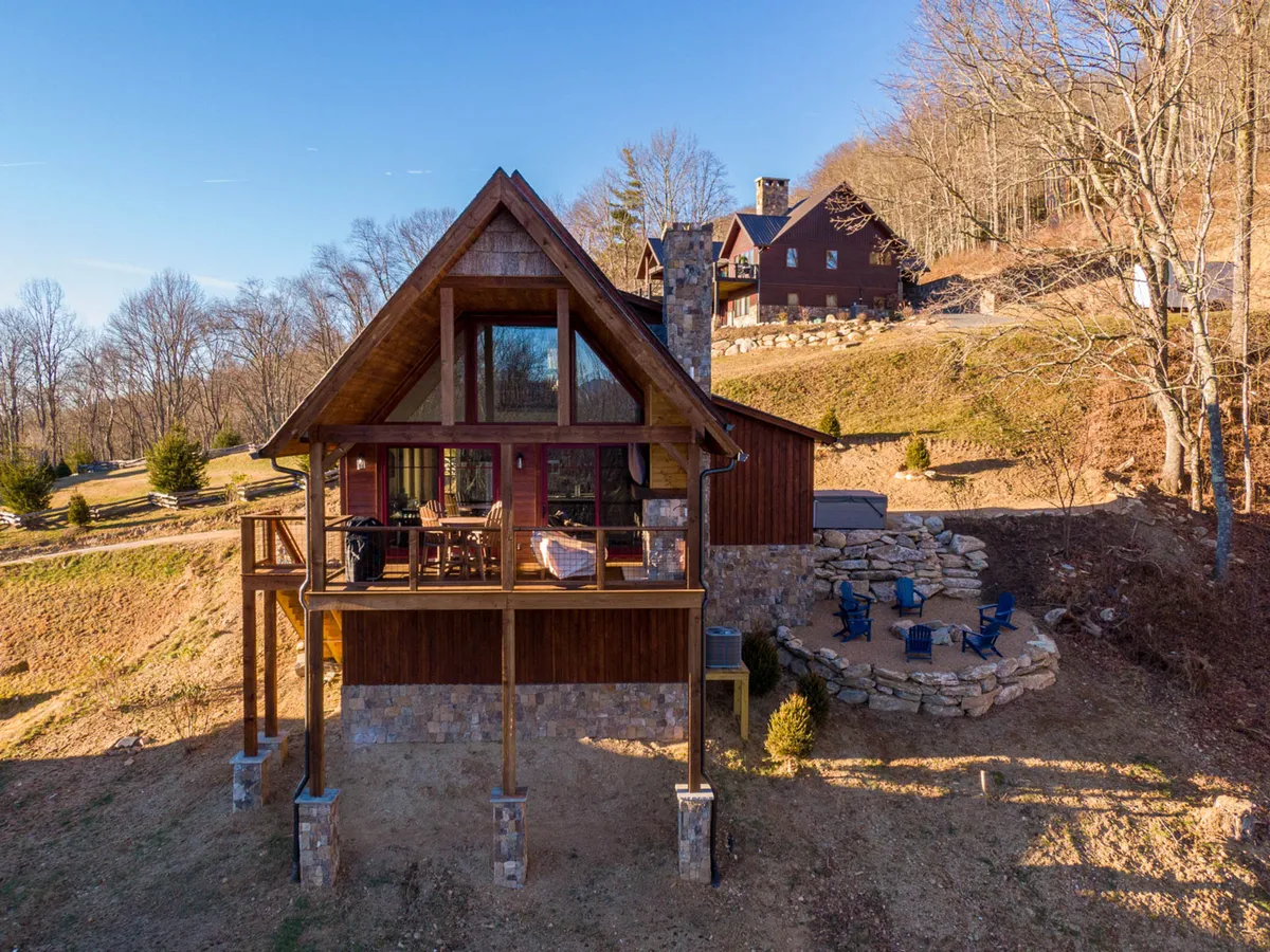 Shows a welcoming exterior of a cabin with an angled roof, large windows, a stone chimney, and an inviting outdoor seating area. The porch offers views ideal for sunsets, stargazing, or morning coffee, with the surrounding mountain landscape creating a serene retreat vibe.
