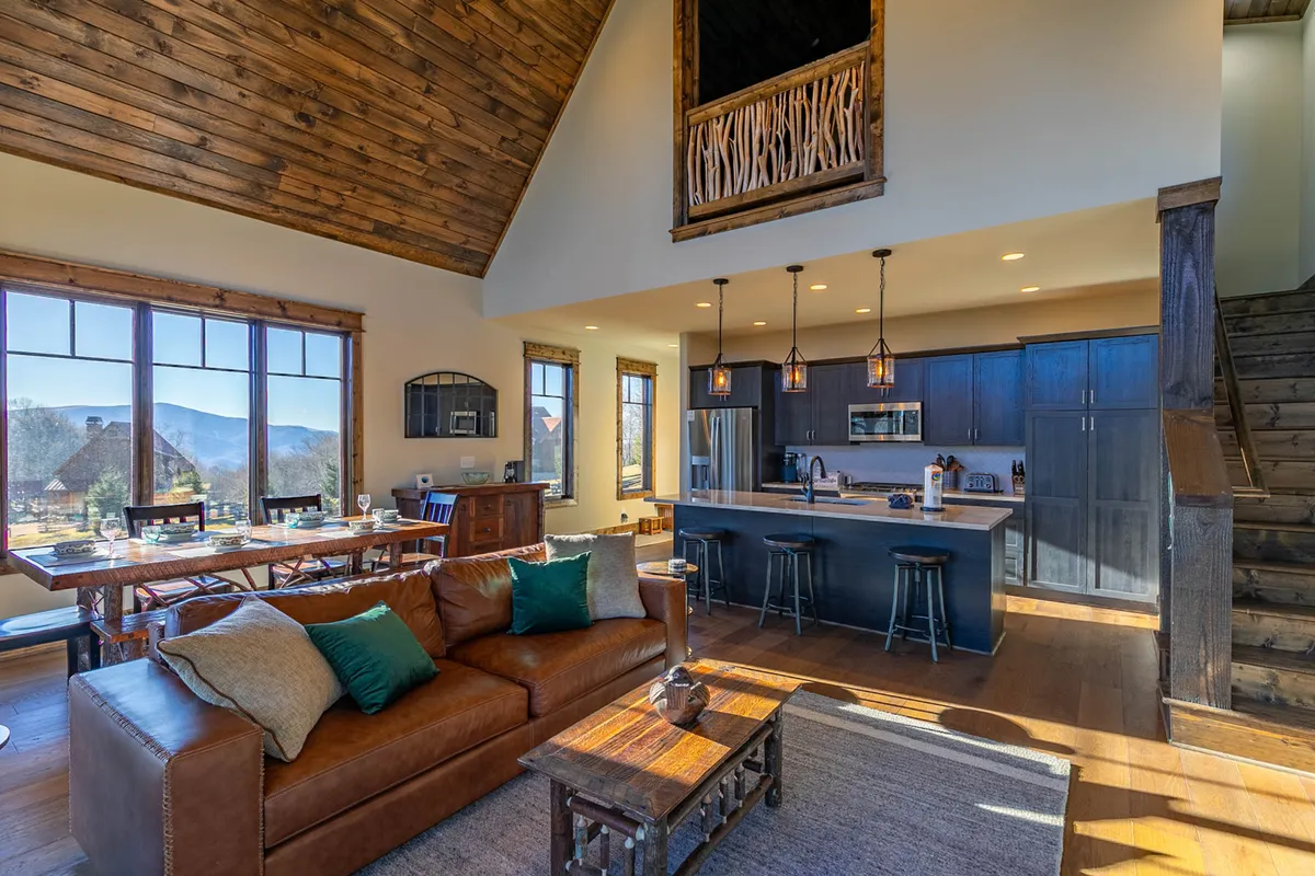 A welcoming open-plan living and dining area featuring a comfortable sofa with colorful cushions and a dining table adjacent to a kitchen island. Large windows fill the space with natural light and reveal glimpses of the outdoors, suggesting a connection to the surrounding environment and potential gathering spaces for community events.