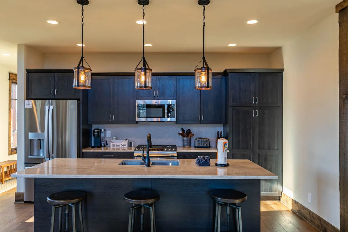 A modern kitchen with a prominent island countertop, hanging pendant lights, sleek stainless steel appliances, and dark cabinetry. The space feels both cozy and functional, perfect for prepping casual snacks or gourmet feasts.