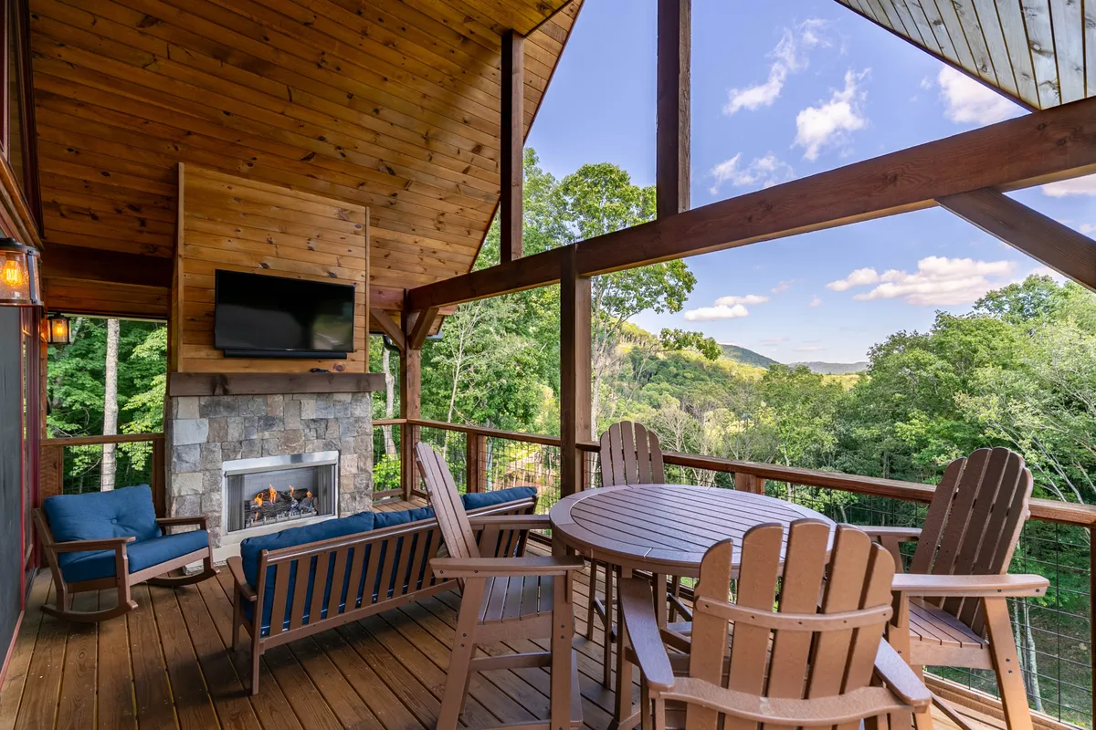 An inviting exterior deck with comfortable seating arranged around an outdoor fireplace, all set against a backdrop of sweeping mountain views. A flat-screen TV is mounted for outdoor movie nights, and the sunset bathes the scene in a cozy, tranquil glow.