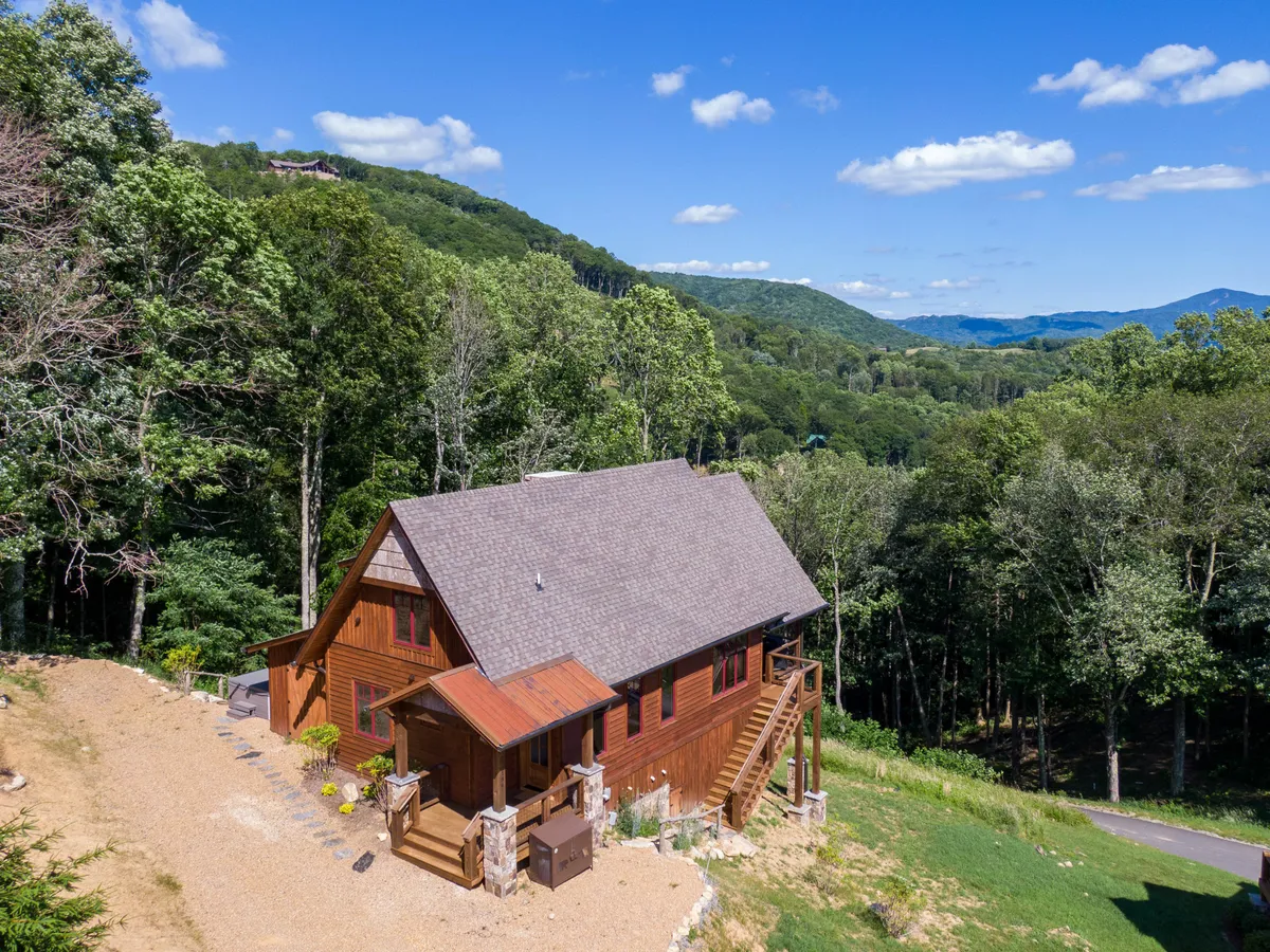This image features a charming wooden cabin nestled among trees, with a view of the mountains in the background and a gravel driveway out front. The porch is visible, suggesting a welcoming space for families to gather. The natural surroundings evoke a sense of tranquility and invite exploration of the outdoors.