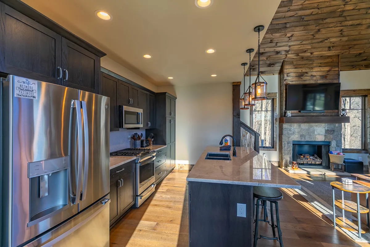 A cozy, modern kitchen scene with stainless steel appliances, a striking granite countertop, and a warm wooden ceiling. The fireplace is visible in the background, complementing the kitchen's inviting atmosphere. The space is designed for both cooking and gathering, with plenty of room to entertain guests or spend quality family time.