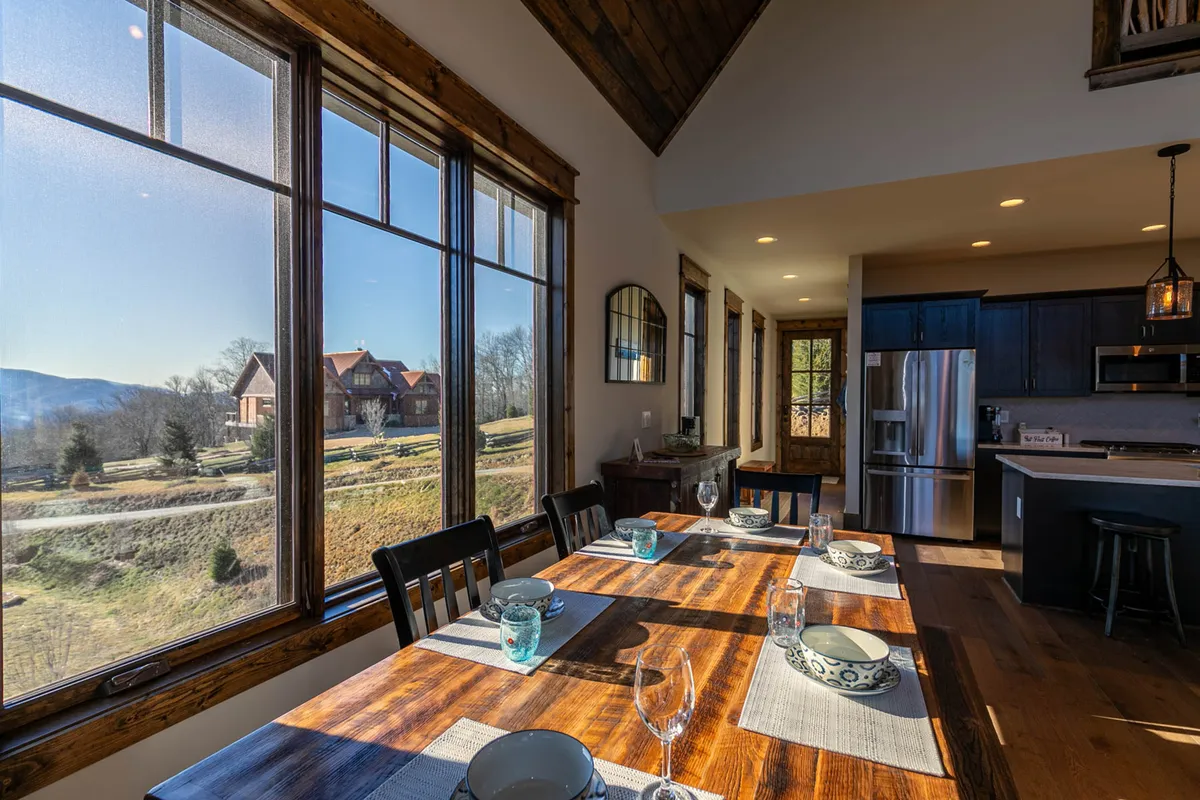 A bright, airy dining area featuring a large table set beside expansive windows that reveal a mountain view. The kitchen is visible in the background, emphasizing the open-concept layout. The setup invites groups to gather for meals while enjoying natural light and scenic vistas.