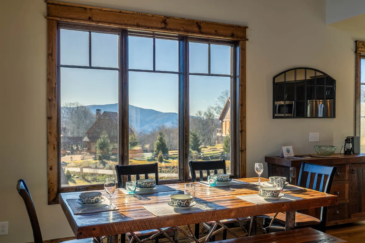This photo focuses on a dining table set beside expansive windows that reveal a sweeping mountain view. Decorative bowls and subtle décor create a warm, welcoming backdrop, while the visible landscape connects the interior space to the surrounding natural beauty.