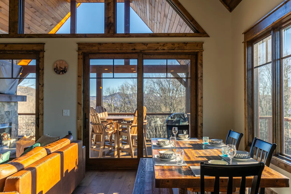 This image captures a spacious dining area with large windows that open out onto a deck furnished with wooden chairs. The scene emphasizes the seamless transition between indoor and outdoor living, with ample natural light streaming in and inviting views just beyond the glass. The setting is cozy and family-friendly, ideal for gatherings or relaxing after a day of adventure.