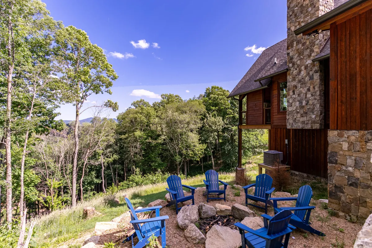 A cozy outdoor space with blue Adirondack chairs arranged around a stone fire pit. The fire pit area is set against the backdrop of the wooden and stone house and is nestled among tall trees, creating an inviting environment ideal for gatherings and storytelling under the stars.