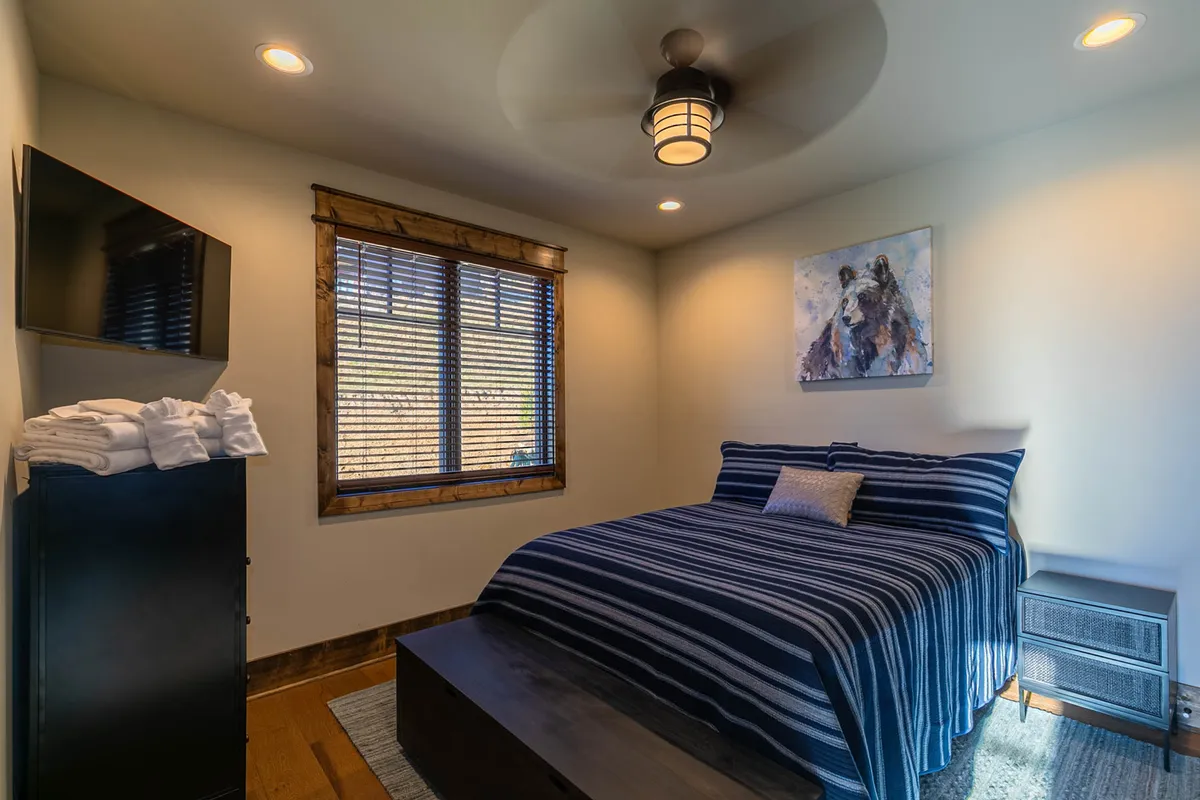 This image shows a peaceful bedroom with a bed dressed in striped bedding, a large window with wooden blinds allowing in natural light, a painting on the wall, and a TV on the dresser. The space feels calm and restful, ideal for relaxation and rejuvenation.