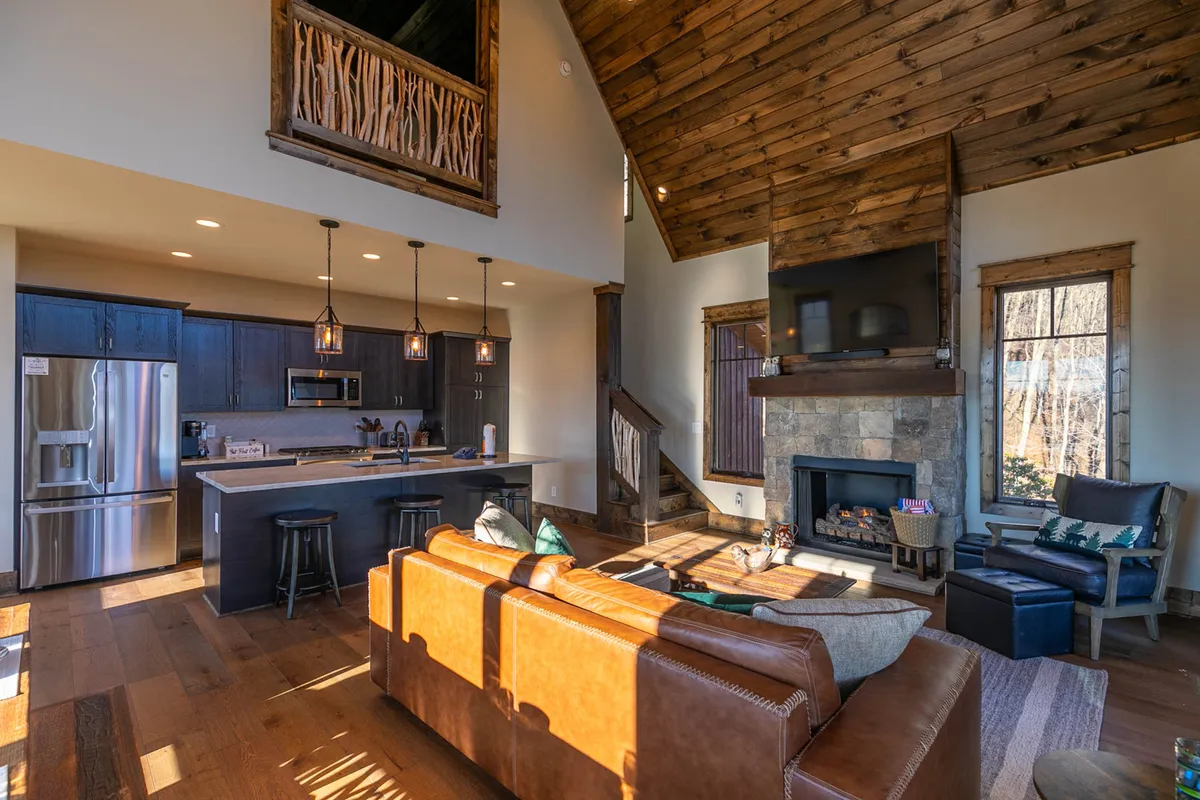 This inviting interior scene features a comfortable sofa, a glowing fireplace, a kitchen island, and dramatic large windows that frame the outdoors. The room is styled for warmth and togetherness, fostering a welcoming atmosphere after a day spent outside.